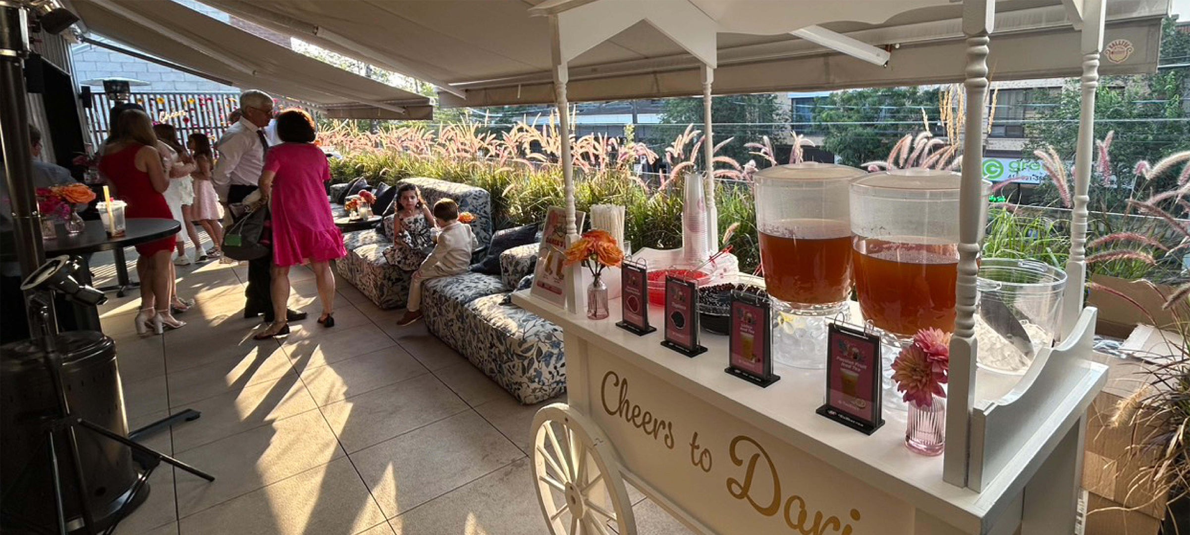 White beverage cart with iced tea dispensers at an outdoor event with guests on a patio